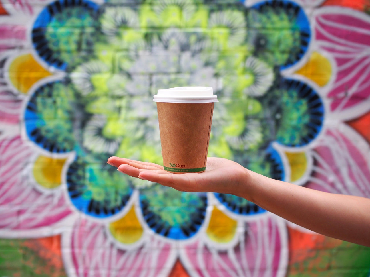 kraft single wall coffee cup with white compostable pla lid being held in front of a graffiti wall