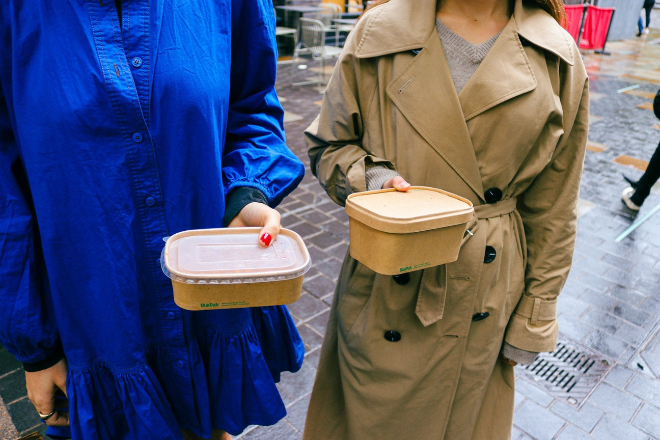 two women holding rectangular kraft takeaway containers with matching kraft and recyclable plastic lids in the rain