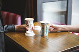 16oz single wall art series coffee cups with sugarcane lids on a cafe table between two friends