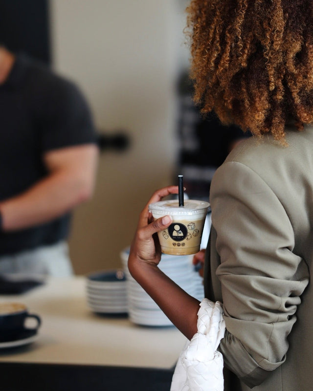 9oz custom branded double sided clear cups with black straw being held by a woman waiting in a coffee shop