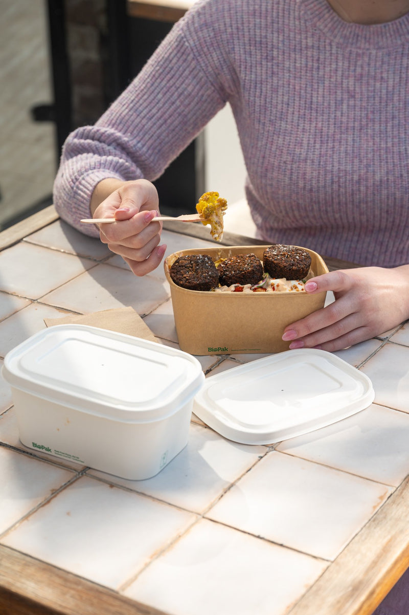 a woman eating a falafel and vegetable takeaway meal from a rectangular kraft food container using wooden cutlery